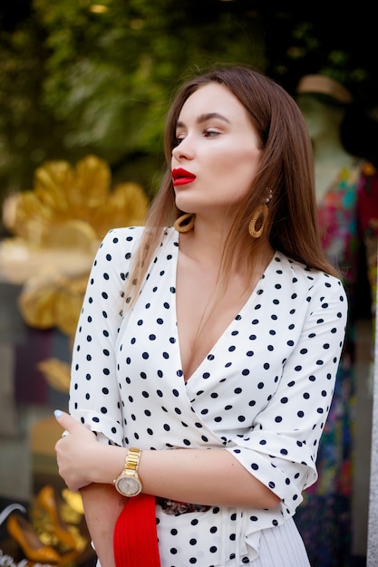 Beautiful model look brunette female wearing white dress with black polka dots is while waking on a city street background with stylish bags in the hand.