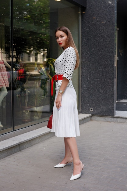 Beautiful model look brunette female wearing white dress with black polka dots is while waking on a city street background with stylish bags in the hand.