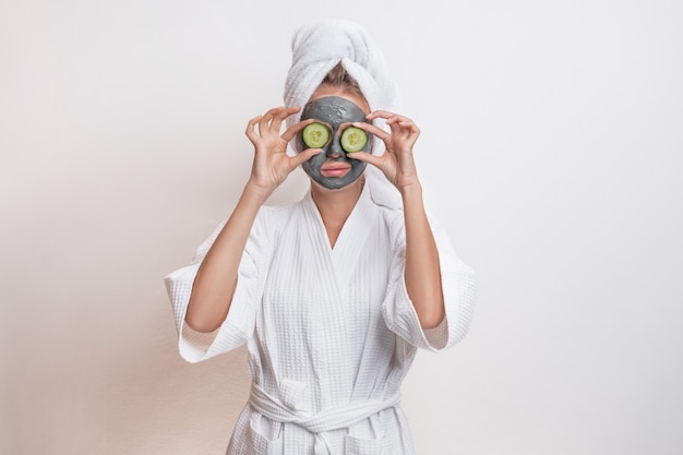 Beautiful  model posing in a bathrobe and a towel on her head holding cucumbers on her eyes with a clay mask on her face 