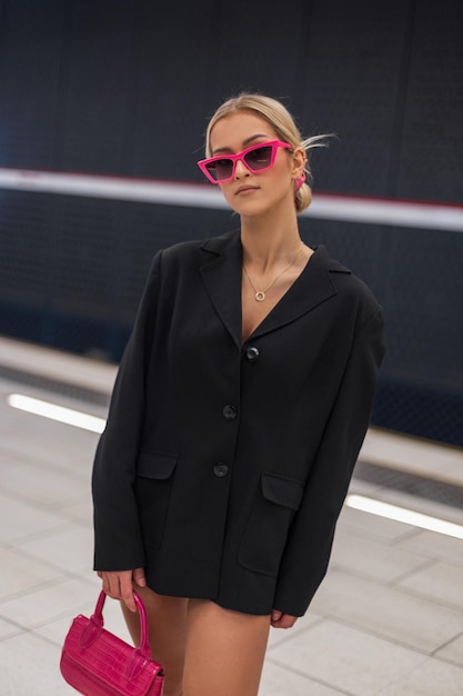 Фото Красивая модель городской женщины в модном черном блейзере с розовыми солнцезащитными очками с розовой сумочкой ходит на станции метро