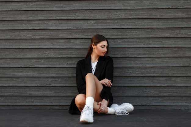 Красивая модель женщина в модном черном пальто с белыми кроссовками сидит у деревянной стены