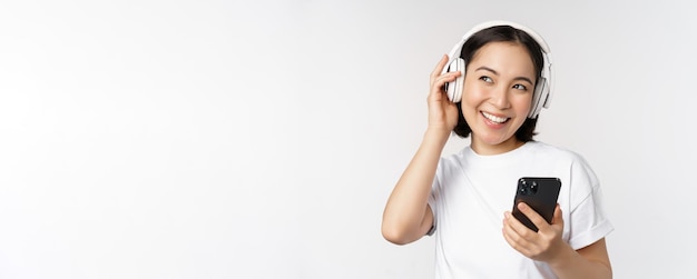 Beautiful modern asian girl listening music in headphones holding mobile phone using smartphone app