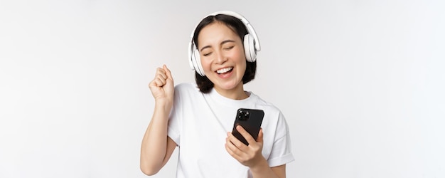 Beautiful modern asian girl listening music in headphones holding mobile phone using smartphone app