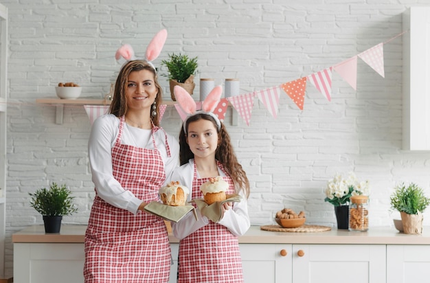 Красивая мама и дочь держат в руках куличи на фоне домашней кухни. Приготовления к празднику Пасхи