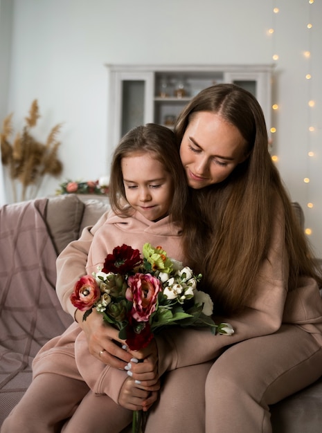 스웨트셔츠를 입은 아름다운 엄마와 딸이 소파에 앉아 꽃으로 어머니의 날을 축하하고 있습니다