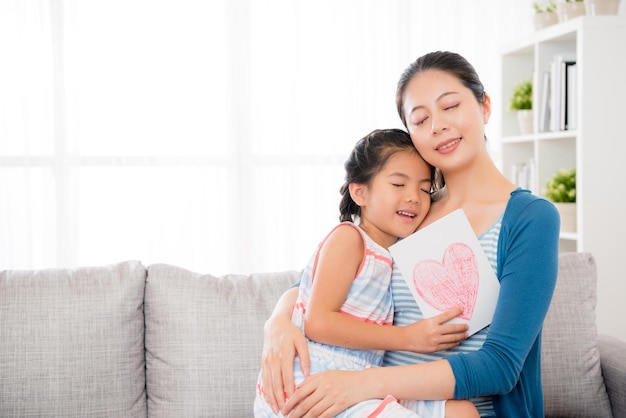 Beautiful mom holding lovely little daughter hug sitting\
together on living room sofa when she receives love card gift happy\
to enjoy mother\'s day holiday at home.