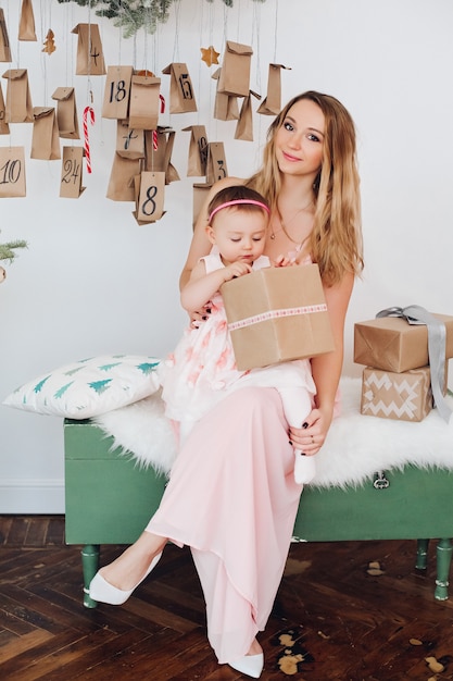 Beautiful mom with little daughter in pink dresses playing near Christmas decoration.