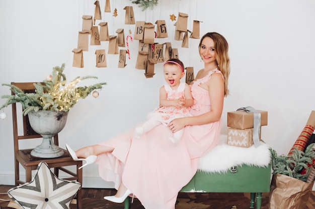 Beautiful mom with little daughter in pink dresses playing near Christmas decoration.