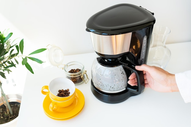 Beautiful morning the girl is waiting for her coffee An automatic drip coffee maker stands with a yellow cup on a white table Electric kitchen small household appliances