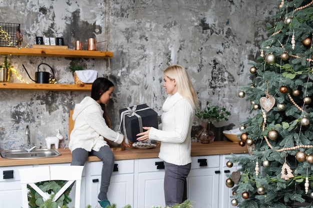 Bella madre e figlia si divertono, si abbracciano e si baciano a casa vicino all'albero di natale in un interno bianco. felicità familiare, vacanza, gioia, vacanza, giochi con una donna. preparativi di capodanno.