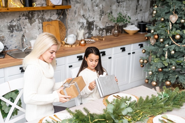 Bella madre e figlia si divertono, si abbracciano e si baciano a casa vicino all'albero di natale in un interno bianco. felicità familiare, vacanza, gioia, vacanza, giochi con una donna. preparativi di capodanno.