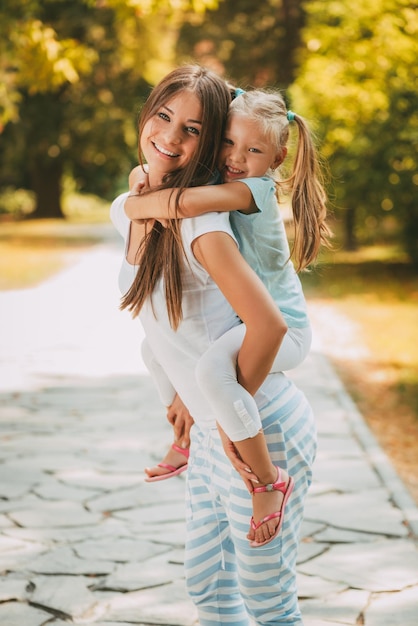 Photo beautiful mother and daughter walking through a park in spring day. young mother piggybacking his daughter.