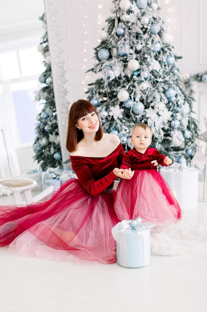 beautiful mother in red dress posing with her cute little baby girl in cozy light decorated studio