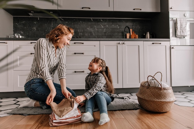 Красивая мать с дочерью, одетые в джинсы и рубашки, веселятся на кухонном полу