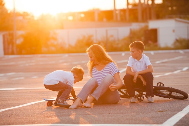 Beautiful mother with her little sons having fun and skating outdoors. She teaches children to ride a bicycle and skateboard