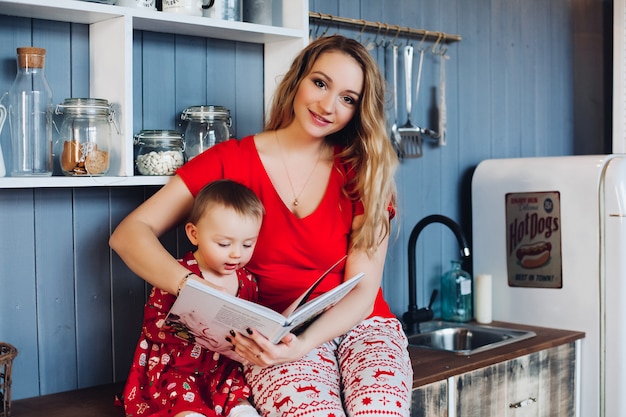 Beautiful mother with little daughter in Christmas pajamas reading book at kitchen.