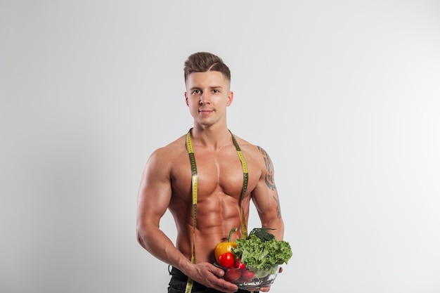 Photo beautiful muscular sports man with a bare torso with a centimeter tape holds a glass bowl of vegetables on a white background in the studio raw healthy food