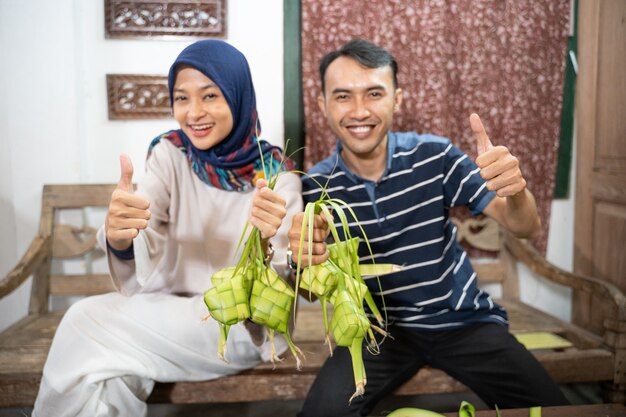 엄지 손가락을 보여주는 eid fitr 무바라크 전통을 위해 야자 잎을 사용하여 집에서 ketupat 떡을 만드는 아름다운 이슬람 가족과 친구