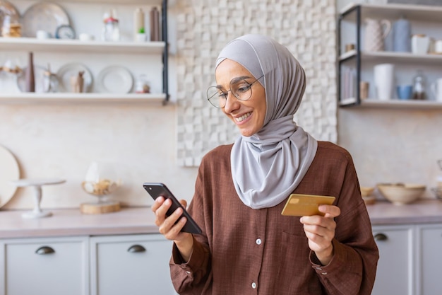 Beautiful muslim girl close up in kitchen woman in hijab smiling and happy holding smartphone and