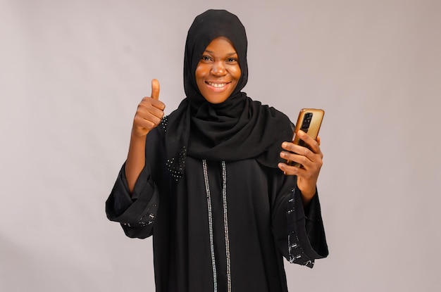 히잡을 쓴 아름다운 이슬람 여성이 휴대전화를 들고 엄지손가락을 치켜들면서 웃고 있다