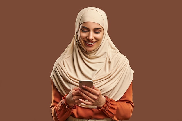 Beautiful Muslim woman in beige hijab holds phone in her hands smiles Advertising technology devices