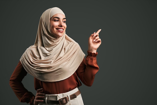 Beautiful muslim woman in beige hijab pointing finger at space on gray background Copy space