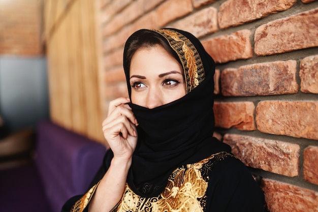 カフェテリアに座っている間、スカーフで顔を覆っている伝統的な摩耗で美しいイスラム教徒の女性。