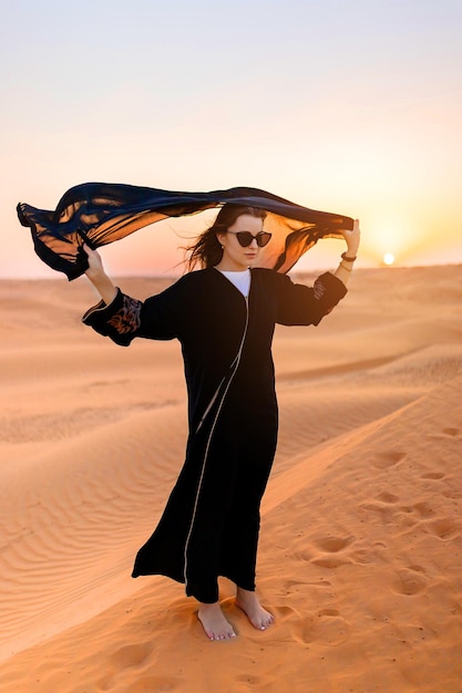 Beautiful mysterious woman in traditional arabic black long dress stands in the desert on sunset