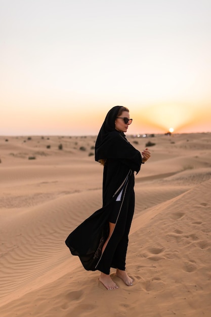 전통적인 아랍어 검은색 긴 드레스를 입은 아름다운 신비한 여성이 일몰에 사막에 서 있습니다.