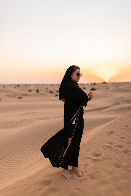 Beautiful mysterious woman in traditional arabic black long dress stands in the desert on sunset