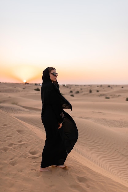 beautiful mysterious woman in traditional arabic black long dress stands in the desert on sunset