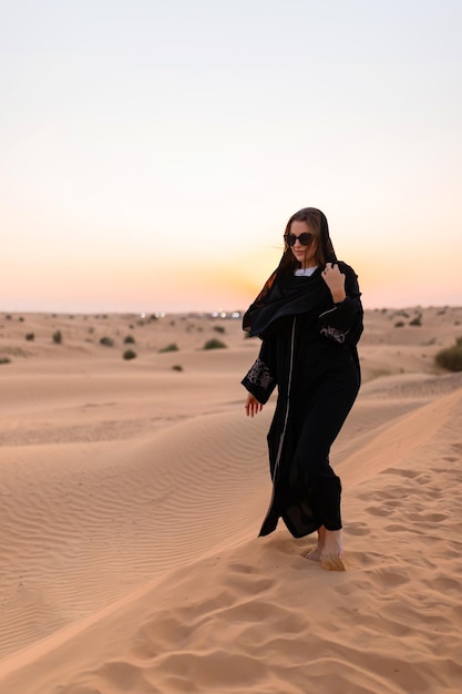 beautiful mysterious woman in traditional arabic black long dress stands in the desert on sunset
