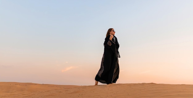 beautiful mysterious woman in traditional arabic black long dress stands in the desert on sunset