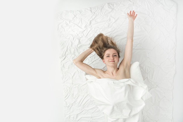 Beautiful naked woman lies in bed hands up, covered with white sheet and enjoys the weekend. Relaxing at home. White background. Top view