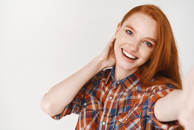 Beautiful and natural girl with ginger hair and blue eyes taking selfie on smartphone smiling lovely at camera front camera view white background