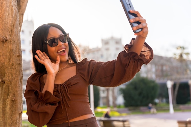 Красивая натуральная молодая африканка в парке в темных очках ведет прямую трансляцию в социальных сетях для подписчиков.
