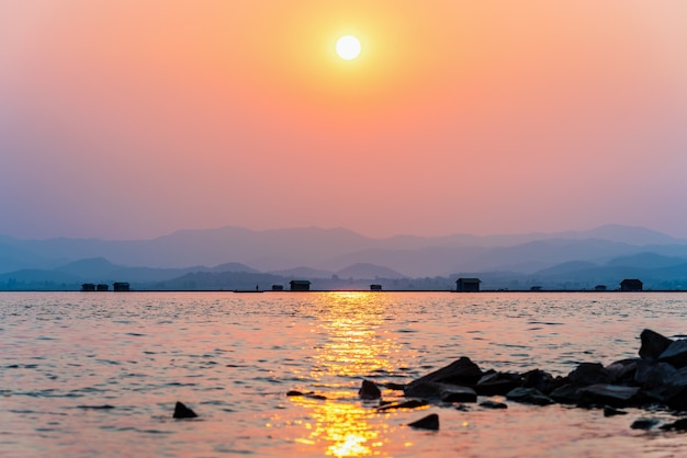 Beautiful nature landscape silhouette fisherman in a boat fishing and fish farming cages yellow sun in the red orange sky at sunset, Local fishing rural lifestyle at Krasiao Dam, Suphan Buri, Thailand