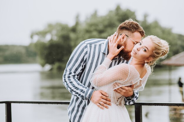 The beautiful newlyweds pose on the pier.The groom gently hugs and kisses the bride's neck on the wharf.