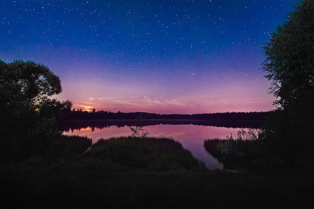 Beautiful night landscape on the forest lake with stars and reflected clouds in water in spring. Colorful sky. Nature background