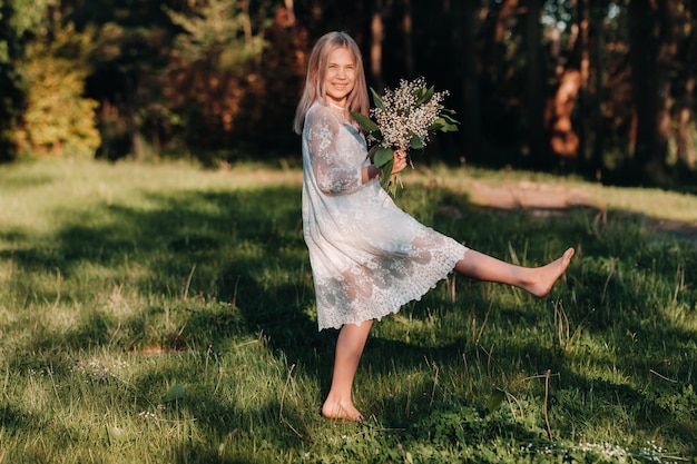 A beautiful nine-year-old blonde girl with long hair in a long white dress, holding a bouquet of lilies of the valley flowers