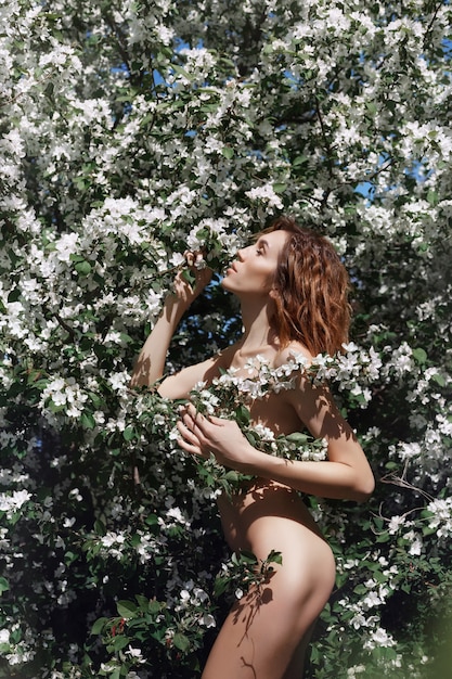 Beautiful Nude art woman in branches and foliage of a flowering Apple tree in nature.