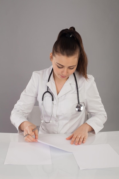Красивая медсестра в халате и со стетоскопом на шее делает отчет за белым столом