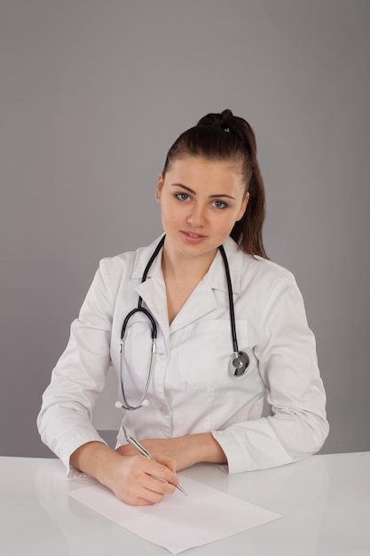 Beautiful nurse in white robe is making her report at the table against of grey background