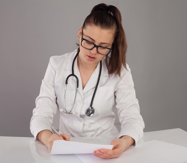 Beautiful nurse in white robe is making her report at the table against of grey background