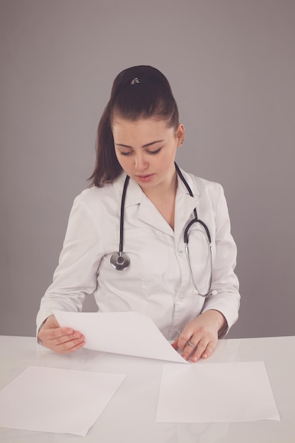 Beautiful nurse in white robe is making her report at the table against of grey background