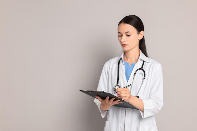 Beautiful nurse with clipboard writing notes on grey background Space for text