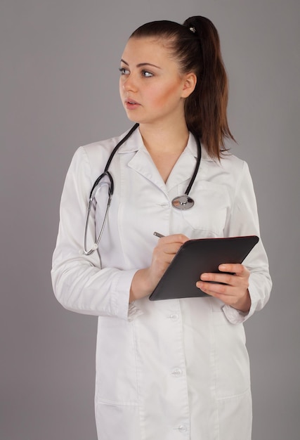 Beautiful nurse with stethoscope is writting something against of grey background