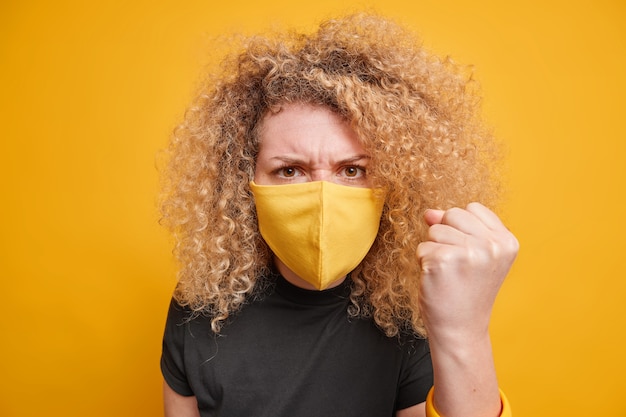 Beautiful outraged young woman with curly hair clenches fist feels very angry expresses negative emotions wears protective face mask during coronavirus pandemic isolated over yellow wall