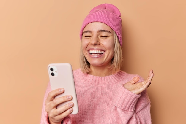 Beautiful overjoyed woman has video call via smartphone laughs out happily discusses something funny keeps palm raised closes eyes dressed casually poses against brown background. Happy conversation