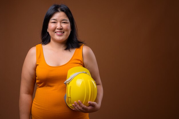 beautiful overweight Asian woman construction worker holding hardhat on brown